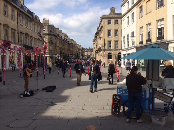 A busker playing on Bath High Street, with some pedestrians and and ice cream stand