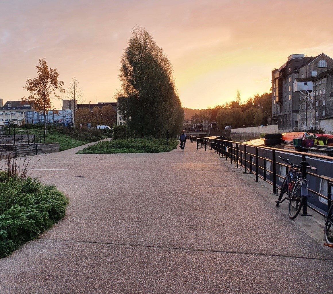 A canal side paved area, with boats moored on the canal, and someone cycling along the pavement at sunset