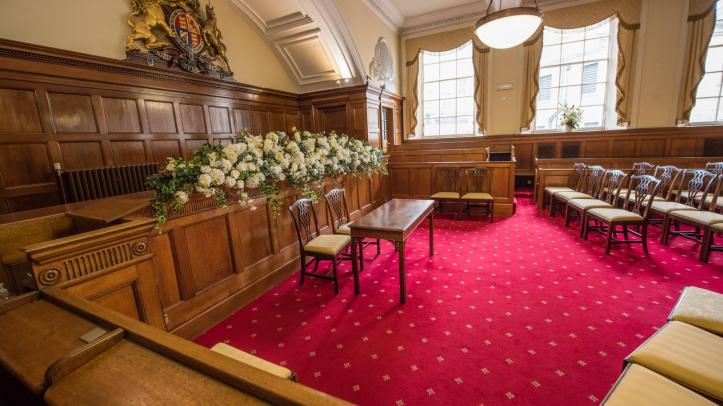 A wooden panelled room with a plush red carpet laid with wooden chairs arranged around a floral display