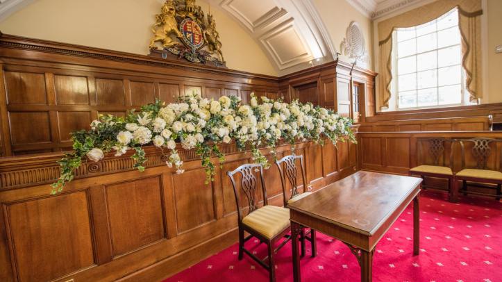 A wooden panelled room with a plush red carpet. Two wooden chairs are behind a wooden table in front of wood panelling and a spectacular arrangement of flowers