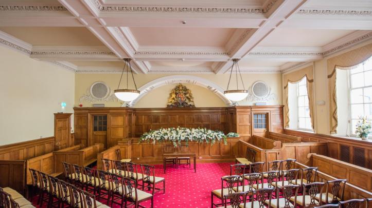 A wooden panelled room with a plush red carpet laid with wooden chairs arranged around a floral display